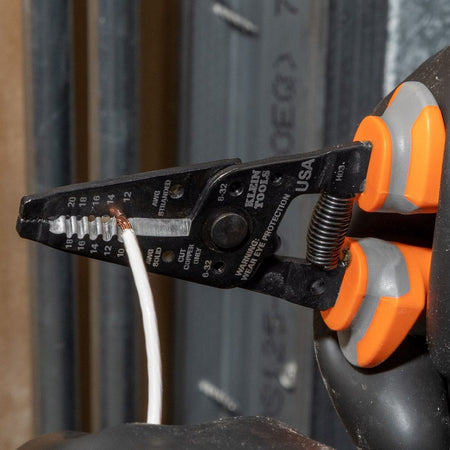 A close-up field action view of an operator using a black-oxide Klein Tools wire stripper cutter from the 1000V insulated hand tool set to strip a white electrical cable, displaying the precision ground stripping holes and spring-loaded handle connection equipped with dual-component orange and gray thumb guards.
