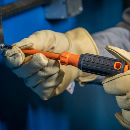 A close-up view of a technician wearing heavy-duty tan leather safety gloves holding an orange Klein Tools insulated screwdriver, showcasing the flame-retardant orange insulation running flush down to the black alloy drive tip face as it turns a metal screw fastener.
