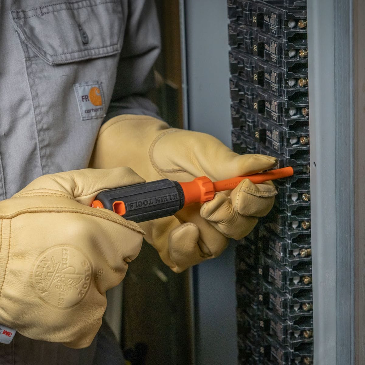 An electrician wearing durable, stamped tan leather safety work gloves uses a 1000V insulated cabinet tip screwdriver to tighten a screw terminal inside a commercial circuit breaker panel box, highlighting the ergonomic black rubberized cushion-grip handle and flame-retardant orange dielectric shank insulation.