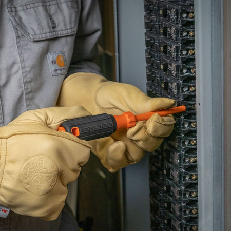 An electrician wearing durable, stamped tan leather safety work gloves uses a 1000V insulated cabinet tip screwdriver to tighten a screw terminal inside a commercial circuit breaker panel box, highlighting the ergonomic black rubberized cushion-grip handle and flame-retardant orange dielectric shank insulation.