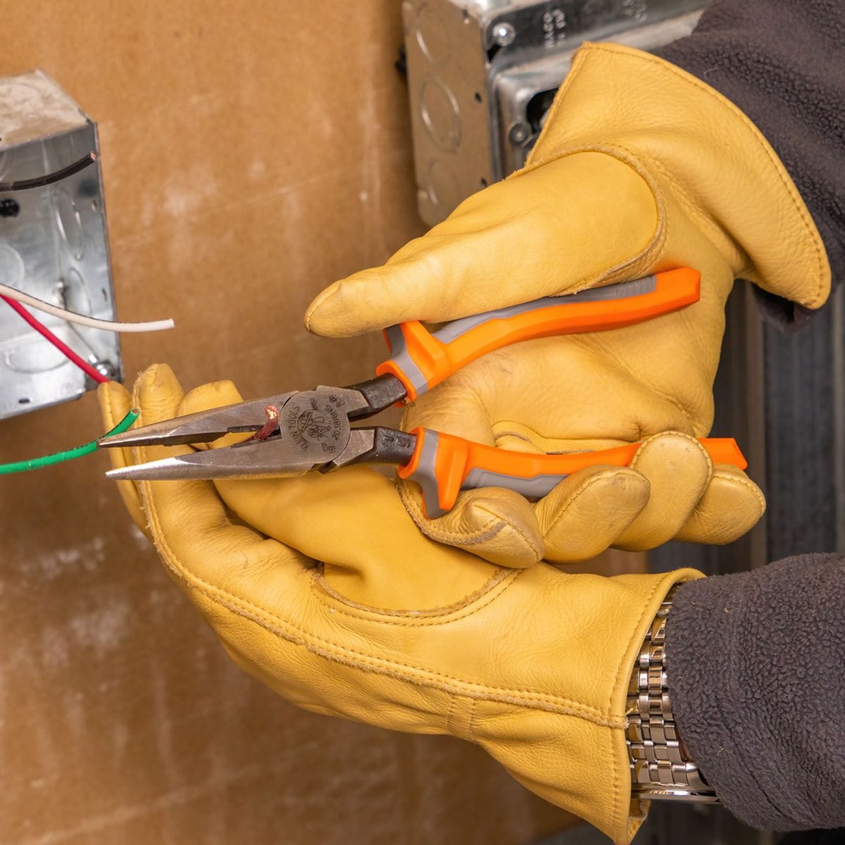 An electrician wearing heavy tan leather work gloves holds a pair of Klein Tools 1000V insulated long-nose pliers to cut a copper wire loop coming from a metal electrical outlet box box, showcasing hand protection and precision alignment near terminal zones.