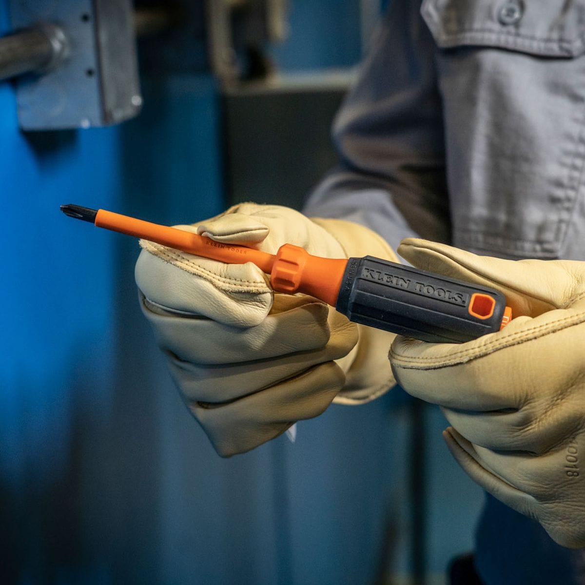A close-up field view of an operator wearing heavy-duty tan leather safety gloves holding a Klein Tools Phillips insulated screwdriver, showcasing the flame-retardant orange insulation running flush down to the black alloy drive tip face, engineered to eliminate flashovers in tight distribution blocks.