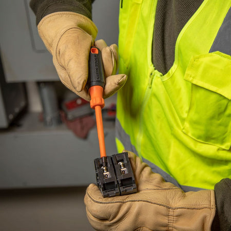 Close-up action shot of a trade professional wearing a high-visibility yellow vest and leather safety gloves using an orange insulated square-drive screwdriver from the 5-piece heavy-duty set to secure a wire lead into a loose black terminal block component.