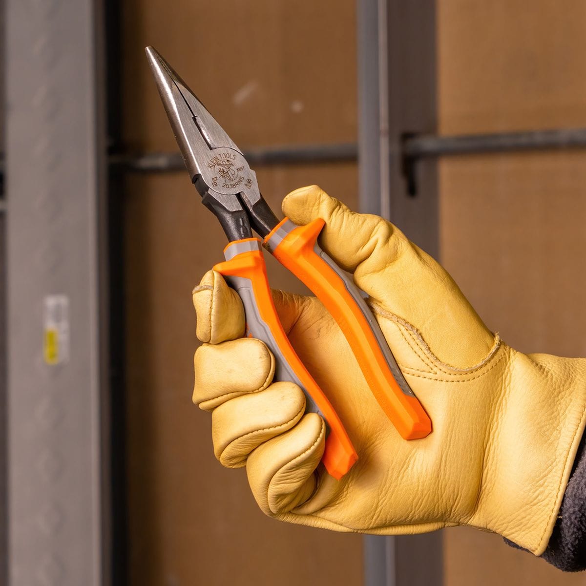 A close-up real-world shot of a trade professional wearing tan leather gloves holding a pair of Klein Tools 2038RINS insulated long-nose pliers, demonstrating the safety handling guard barriers and orange and gray non-slip shock protection handles.