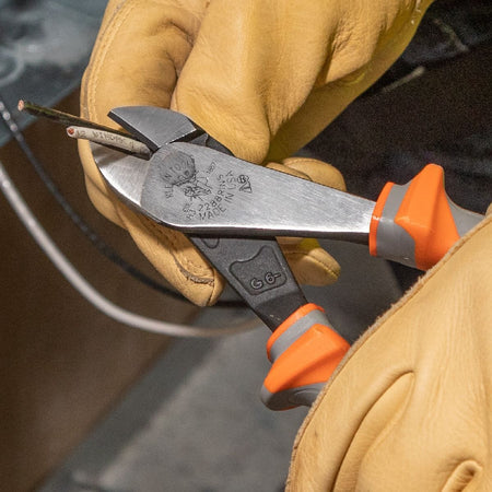 An electrician wearing durable tan leather safety work gloves uses a pair of heavy-duty Klein Tools 1000V rated insulated diagonal cutting pliers to cleanly slice through a multi-conductor electrical wire core, demonstrating the heavy leverage cutting jaw action and non-slip protective orange and gray thumb guard handle profile on a jobsite.