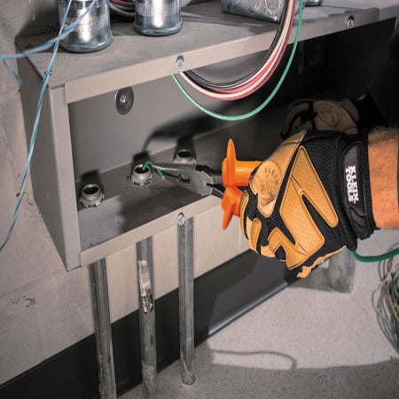 An electrician wearing heavy duty tan leather work safety gloves uses a pair of Klein Tools 1000V insulated long nose pliers to reach inside a gray metal distribution wire box enclosure to route an insulated green ground wire, demonstrating industrial diagnostic utility.