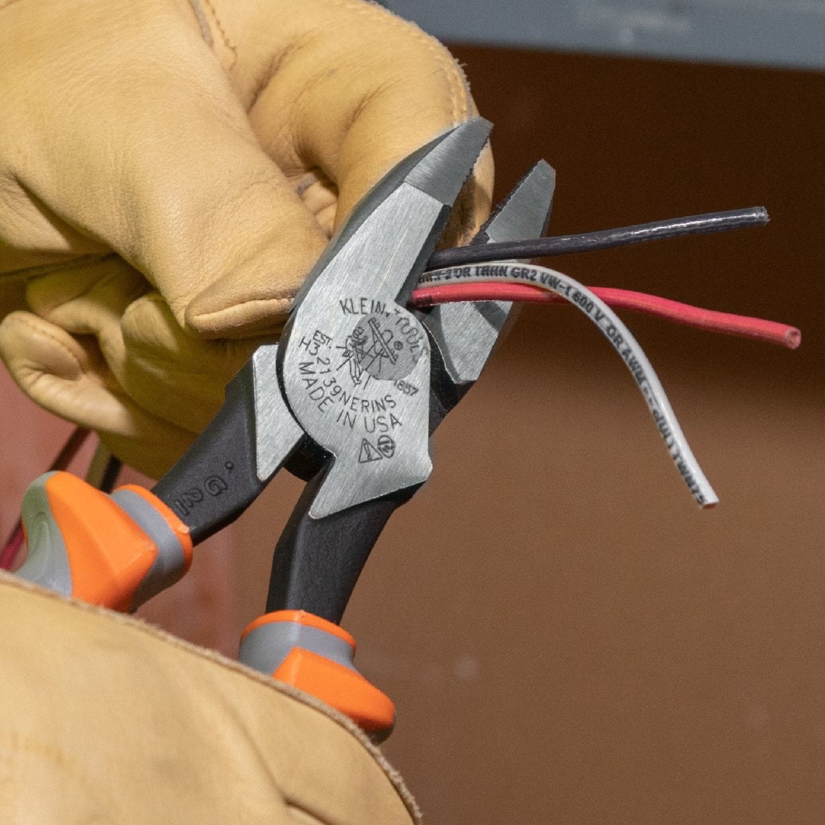 An electrician utilizing a pair of heavy duty Klein Tools side cutting lineman pliers to slice clean lines through three multi colored power wires, demonstrating the cutting force of the induction hardened blades while protected by high dielectric gray and orange handle guards.