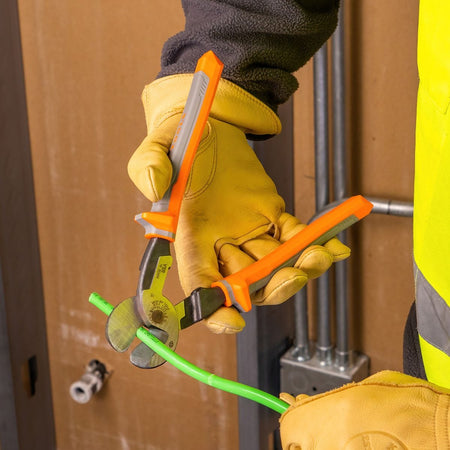 A technician utilizing the high-leverage insulated shear blades of a pair of Klein Tools cable cutters to slice cleanly through a thick green plastic-jacketed copper electrical power supply cable, demonstrating hot-line safety procedures and commercial trade application compliance.