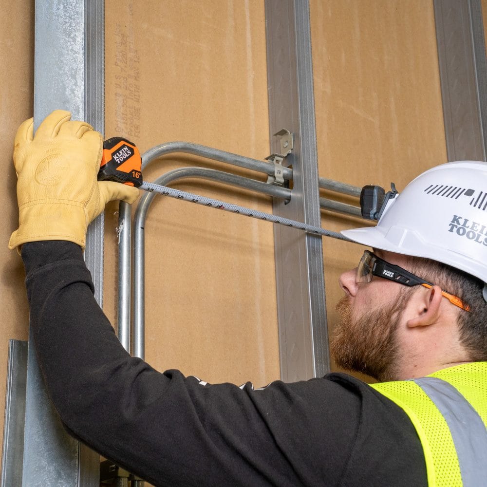 An electrician using the Klein Tools 16-foot tape measure to check the alignment of conduit pipes on a metal stud wall.