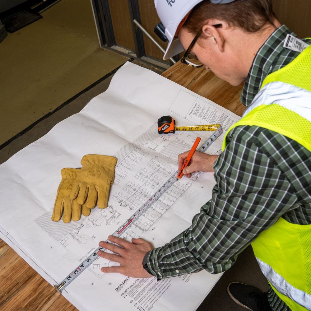 An architect or contractor using a Klein Tools tape measure and orange pencil to verify dimensions on a large set of building plans.
