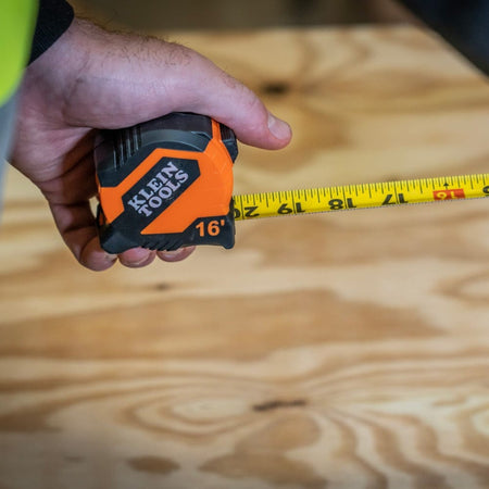 A close-up view of a professional's hand holding the Klein Tools compact tape measure while measuring a wooden surface.