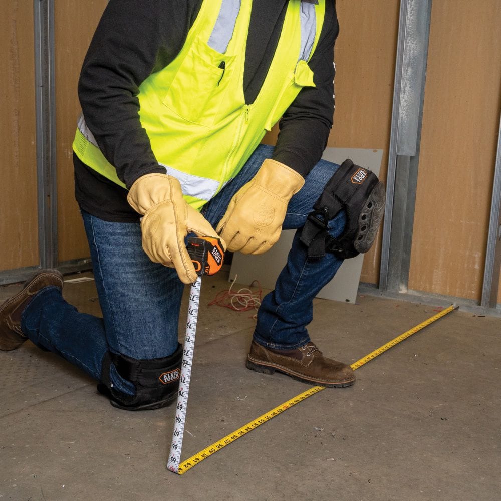 A professional tradesman in safety gear using the double-sided scale of the Klein Tools tape measure to take precise floor-to-wall measurements.