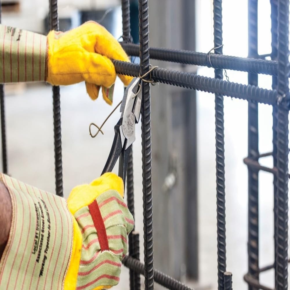 Action shot showing a construction worker with yellow safety gloves using the ironworker's pliers to twist wire ties onto a vertical rebar cage.