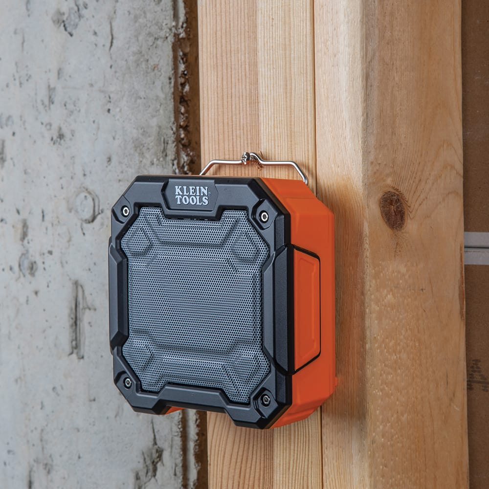 Klein Tools AEPJS3 rugged portable Bluetooth jobsite speaker hanging from a screw on a wooden stud using its integrated metal wire hook.
