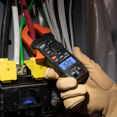 Close-up of a professional electrician using the Klein Tools CL120 Digital Clamp Meter to measure current on a red wire inside a service panel. The backlit LCD clearly displays 2.0 Amps, showcasing the tool's precision and visibility in dark electrical environments.