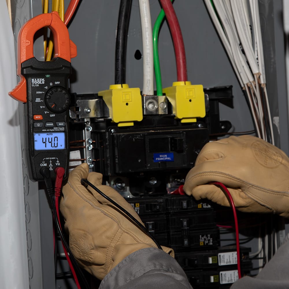 Action shot of the CL120 Digital Clamp Meter being used to troubleshoot an electrical panel. The meter is shown attached to a conductor with the test leads probing circuit breakers, while the high-contrast display indicates an auto-ranging measurement of 44.0 Amps.