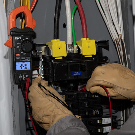 Action shot of the CL120 Digital Clamp Meter being used to troubleshoot an electrical panel. The meter is shown attached to a conductor with the test leads probing circuit breakers, while the high-contrast display indicates an auto-ranging measurement of 44.0 Amps.
