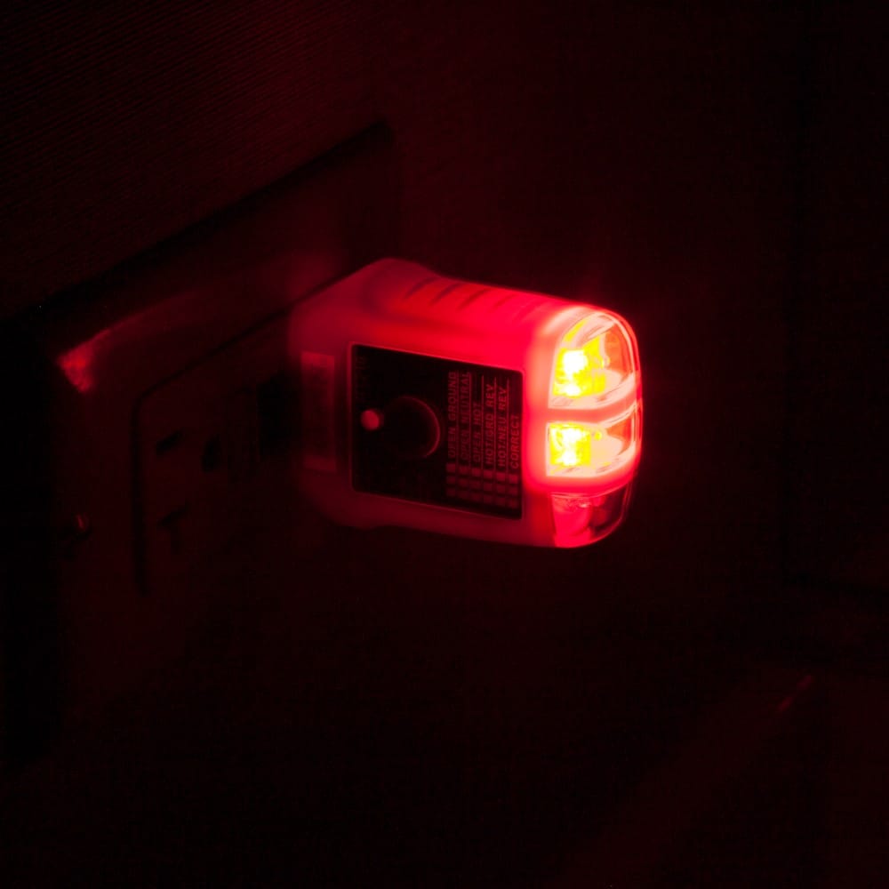 The Klein Tools RT210 GFCI Receptacle Tester plugged into a wall outlet in a dark room. The bright orange visual indicators are illuminated, demonstrating the high-visibility LED design that allows for easy wiring fault identification in low-light conditions.