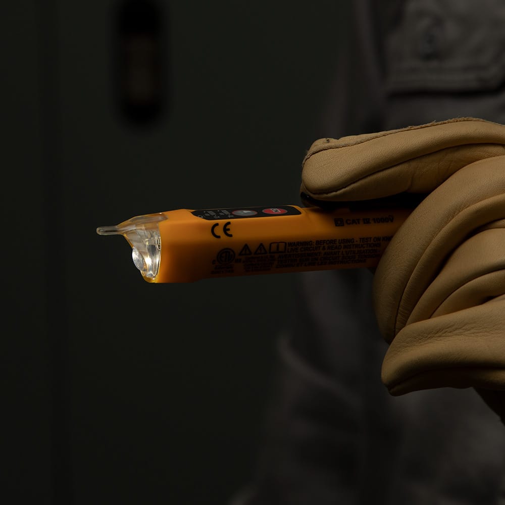 Close-up of the Klein Tools NCVT-3P voltage tester held in a gloved hand with the integrated high-intensity flashlight turned on. The focused beam illustrates the tool's ability to light up dark workspaces independently of the non-contact voltage detection function.