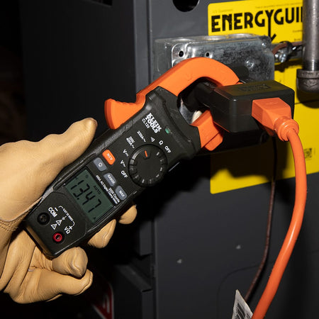 The CL120 clamp meter used to measure current flow through an orange extension cord plugged into a heavy-duty adapter. The technician monitors the display, which shows 13.47 Amps, demonstrating the meter's utility for checking load requirements on standard power equipment.