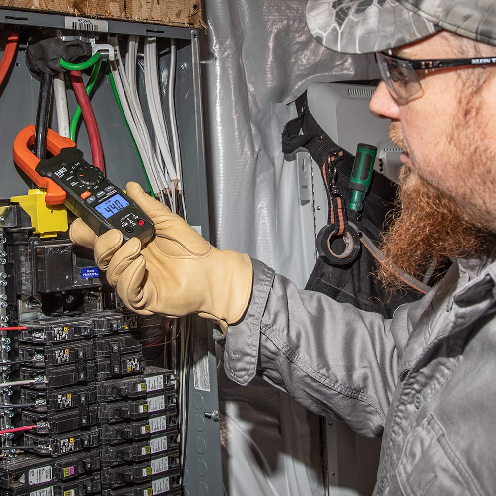 An electrician wearing a hard hat, safety glasses, and protective leather work gloves uses the Klein Tools CL120 Digital Clamp Meter to measure AC current in a residential breaker panel. The meter's orange clamp is securely looped around a main conductor, and the backlit LCD screen clearly displays a measurement of 44.0 Amps, demonstrating the tool's effectiveness for live circuit diagnostics.