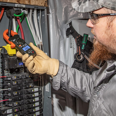 An electrician wearing a hard hat, safety glasses, and protective leather work gloves uses the Klein Tools CL120 Digital Clamp Meter to measure AC current in a residential breaker panel. The meter's orange clamp is securely looped around a main conductor, and the backlit LCD screen clearly displays a measurement of 44.0 Amps, demonstrating the tool's effectiveness for live circuit diagnostics.