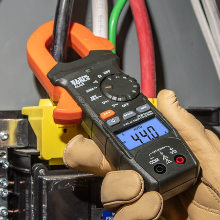 Close-up of the Klein Tools CL120 being used by an electrician to measure current in an electrical panel. The orange clamp is securely placed around a single black conductor, and the backlit LCD displays a clear reading of 44.0 Amps.