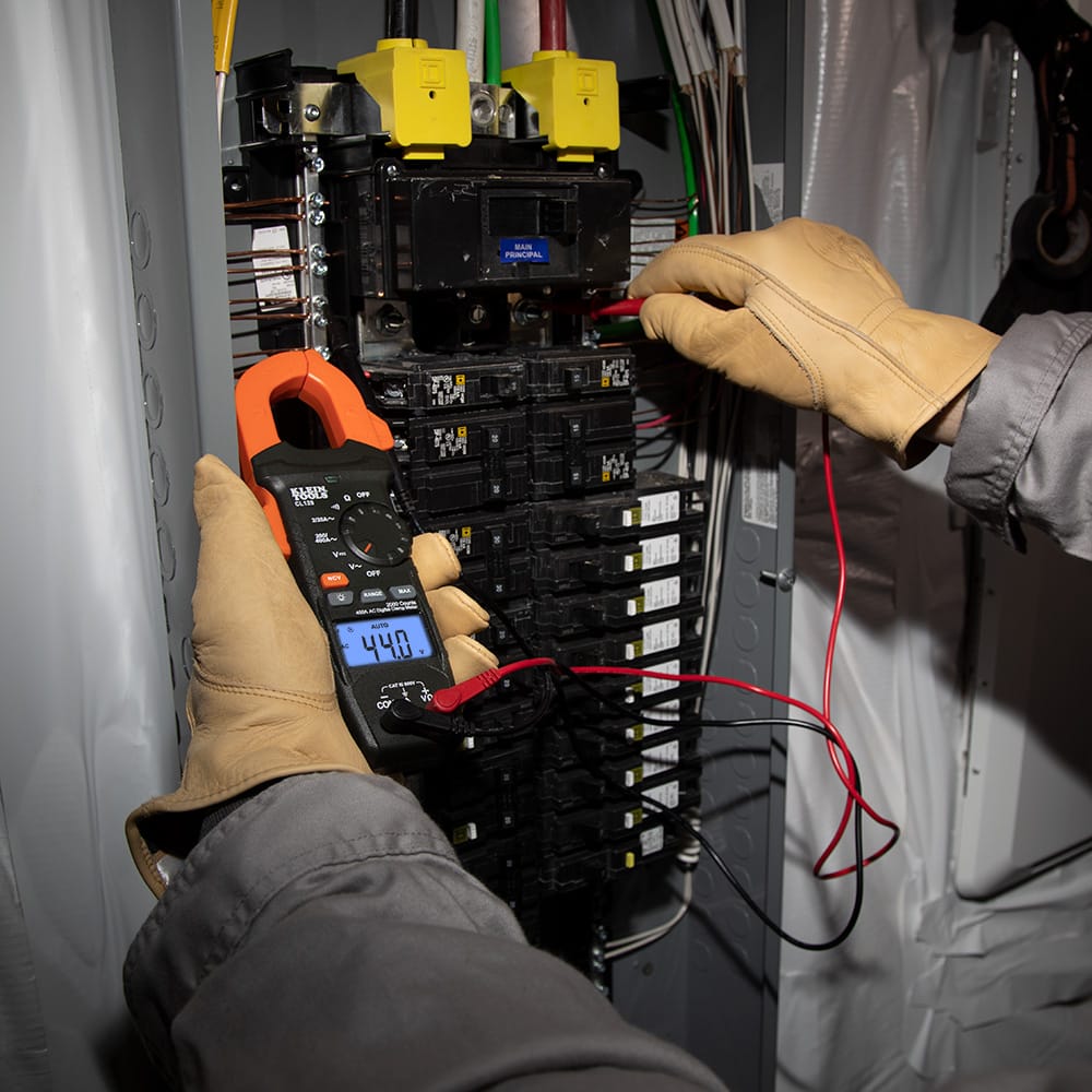 An electrician wearing protective leather gloves uses the CL120 clamp meter and test leads simultaneously to troubleshoot a commercial breaker panel. The backlit display shows $44.0V$, demonstrating the tool's ability to provide clear readings in high-density electrical environments.