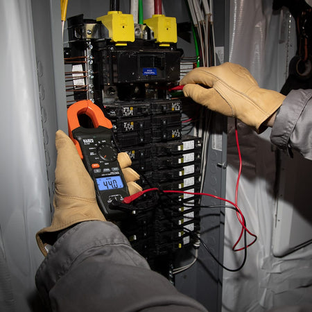 An electrician wearing protective leather gloves uses the CL120 clamp meter and test leads simultaneously to troubleshoot a commercial breaker panel. The backlit display shows $44.0V$, demonstrating the tool's ability to provide clear readings in high-density electrical environments.
