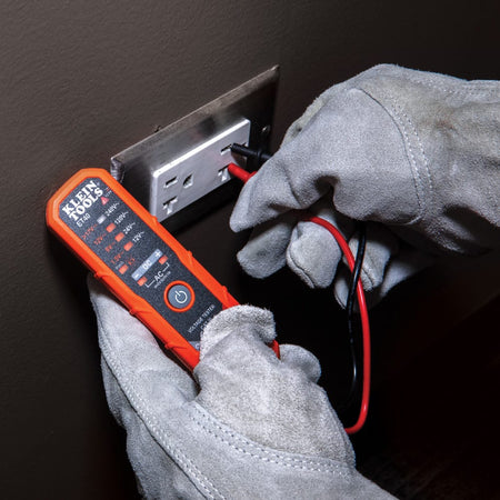 A lifestyle shot of a technician wearing heavy-duty gray suede protective gloves using the Klein Tools ET40 electronic voltage tester. The professional is inserting the red and black test probes into a standard wall outlet to verify the live AC voltage levels, which are indicated by the LED display on the orange tester body.