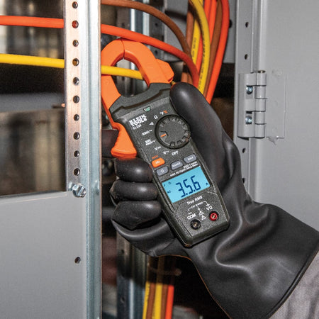 An action shot of an electrician wearing heavy-duty protective gloves using the Klein Tools CL220 clamp meter inside an industrial electrical panel. The backlit LCD screen is clearly visible, displaying a True RMS AC current reading of 35.6 Amps.