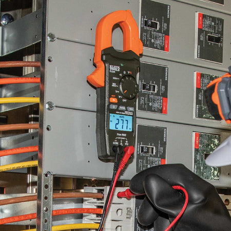 Action shot showing the CL220 being used to verify voltage at a standard electrical outlet. A technician, wearing protective gear, uses the included test leads to monitor real-time AC voltage data, demonstrating the tool's effectiveness for circuit troubleshooting and safety certification.