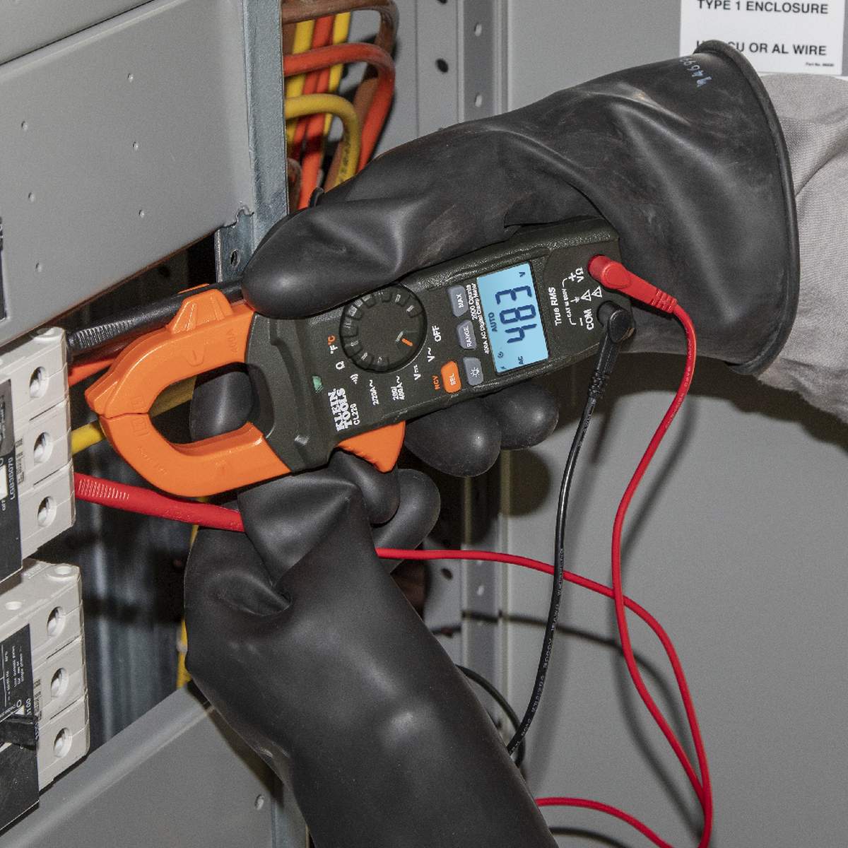 Person wearing black gloves using a digital multimeter with orange clamps on electrical equipment.