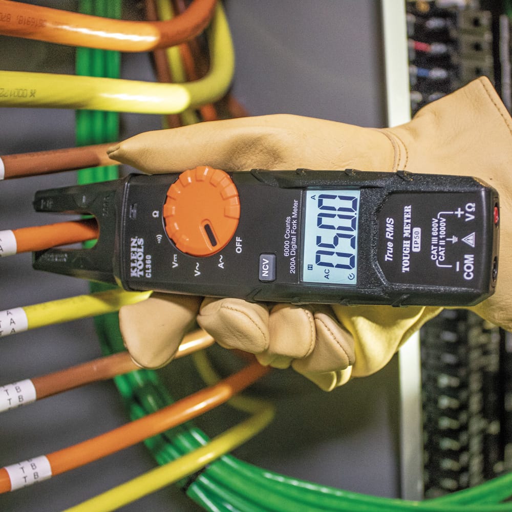 Action shot of the Klein Tools CL360 digital fork meter measuring current in a high-density wiring bundle. The open jaw is positioned around an orange conductor, and the backlit display provides a fast, accurate measurement of 05.00 Amps.