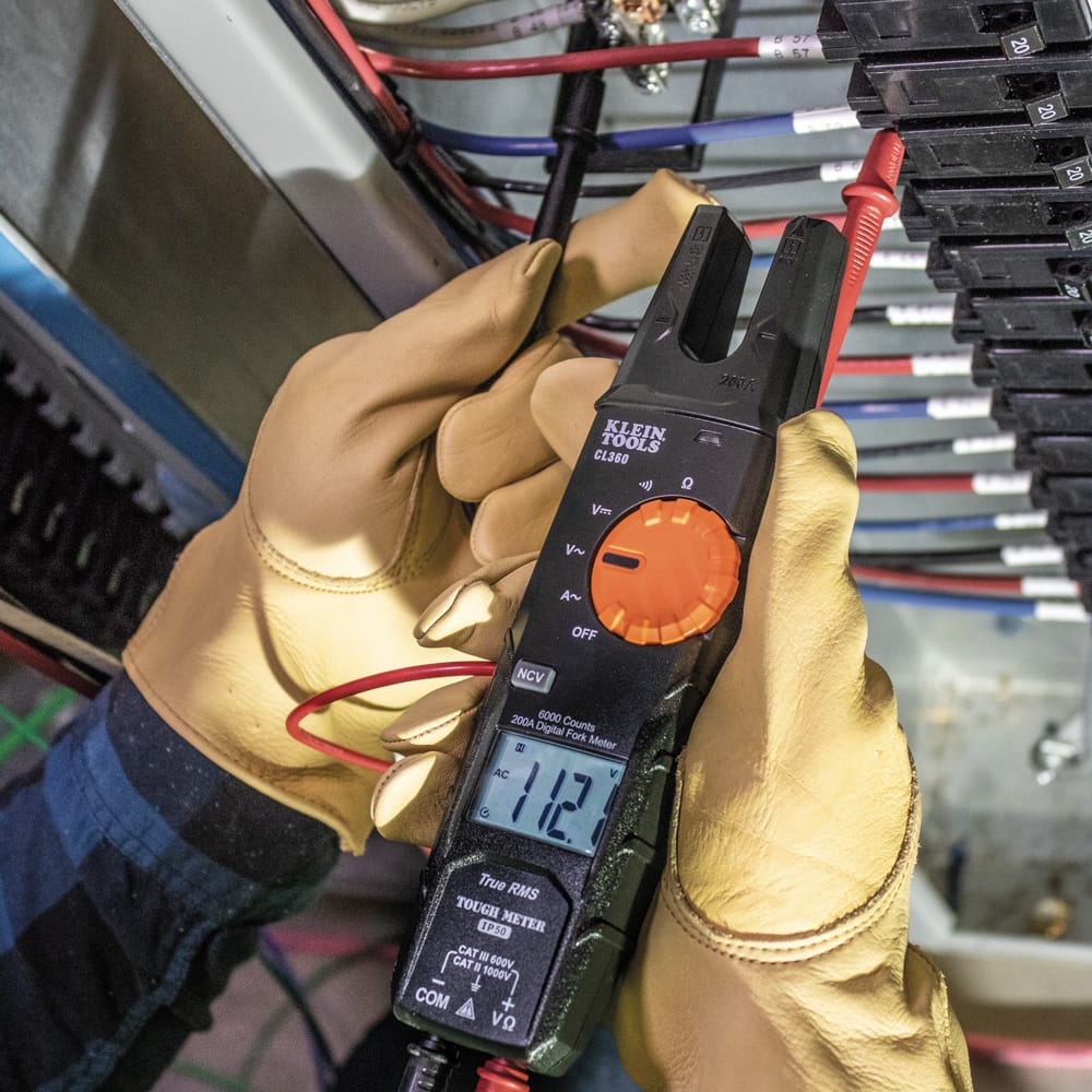 An electrician wearing protective leather gloves uses the CL360 fork meter to measure voltage in a complex industrial panel. The technician uses the test leads to probe the circuit while the meter display shows a clear reading of $11.2V$ AC.