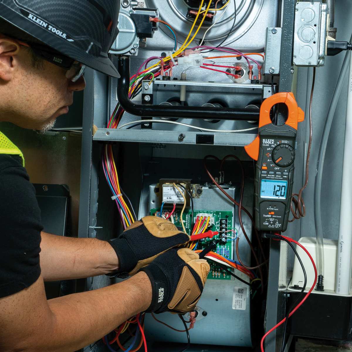 An action shot of a technician using the CL445 to measure AC current in a residential furnace. The clamp jaw is shown encircling a live wire while the integrated worklight illuminates the tight enclosure, providing clear visual feedback on the system's amperage draw.