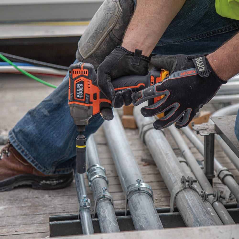 Electrician using a cordless impact wrench with a Klein 1/2 x 3/8-inch coaxial socket to secure conduit clamp hardware on a jobsite floor.