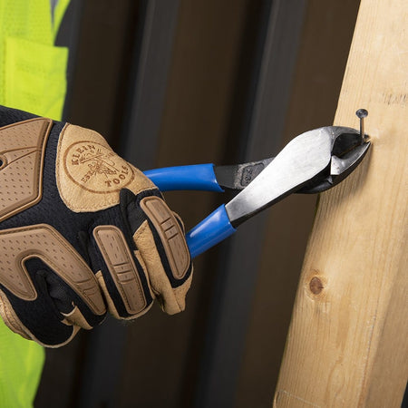 A professional in a high-visibility yellow vest and tan work gloves using the beveled cutting edges of the Klein D2000-48 pliers to easily snip a metal nail embedded in a wooden 2x4 stud.