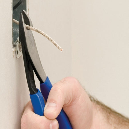 A close-up action photograph of a professional using Klein D2000-48 angled head diagonal cutters to precisely trim a white electrical wire protruding from a wall-mounted metal junction box.