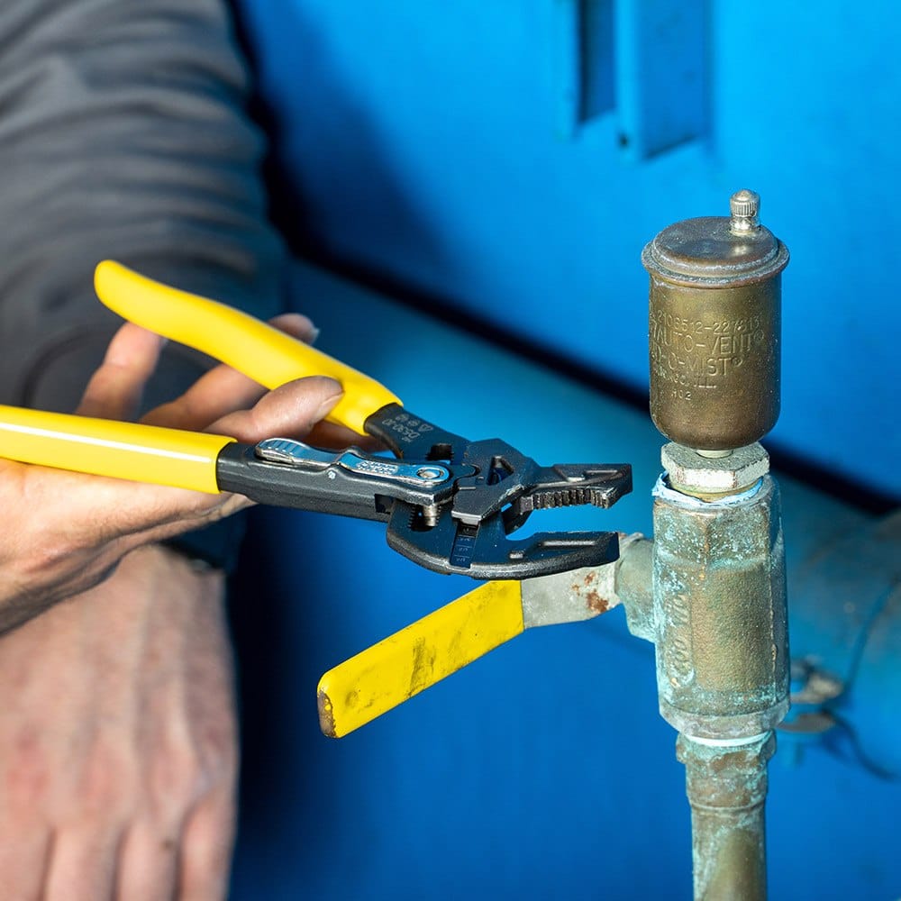 Professional plumber using the Klein Tools adjustable plier wrench to tighten a brass valve on a blue water tank, demonstrating the tool's effectiveness in real-world plumbing applications.