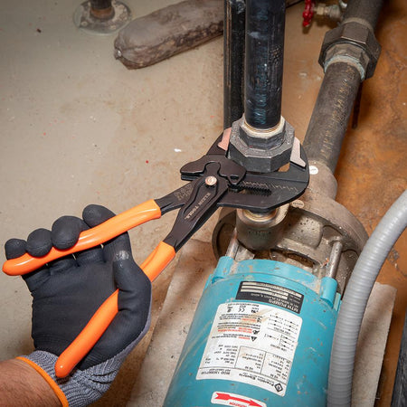 A professional using the 12-inch Klein Tools compact plier wrench to tighten a large hex nut on a blue water pump system, demonstrating high torque and precision.