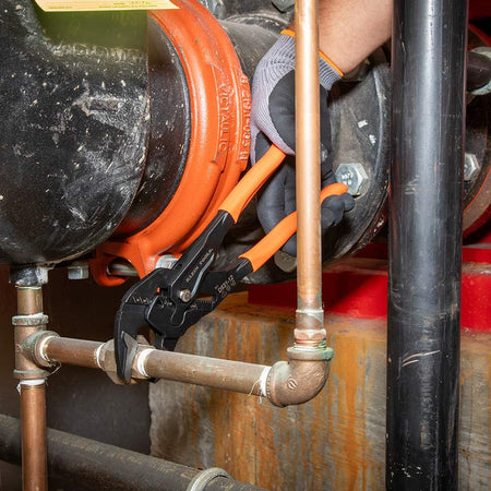 Action shot of a professional using the Klein Tools compact plier wrench to secure a pipe fitting on large industrial machinery, demonstrating its effectiveness in plumbing and mechanical applications.