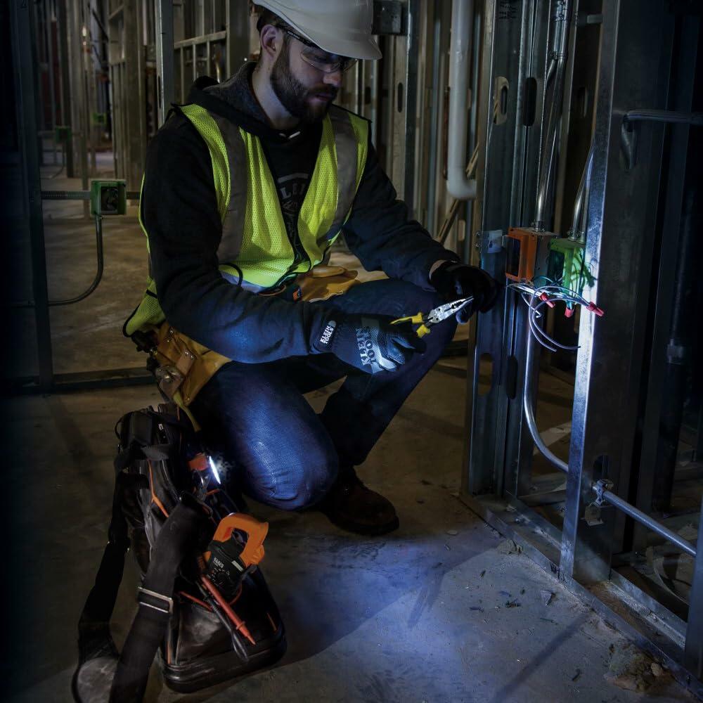Electrician working on wiring inside a metal stud wall, wearing PPE and using tools near a junction box with green, white, and red wires.