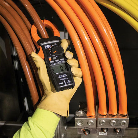 Technician using Klein Tools CL800 clamp meter to measure current on brown cable in a labeled three-phase terminal block (L1, L2, L3).