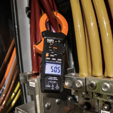 Electrician wearing safety gear using Klein Tools CL800 clamp meter to measure current on color-coded cables inside an electrical panel.