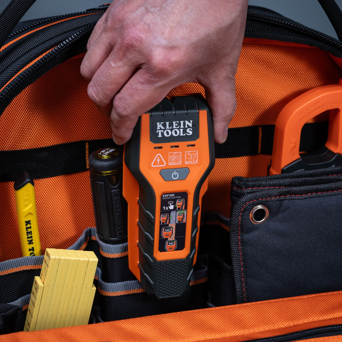 An action shot showing a professional tradesperson’s hand reaching for the compact ESF200 stud finder stored inside a specialized pocket of an orange and black Klein Tools jobsite tool bag.