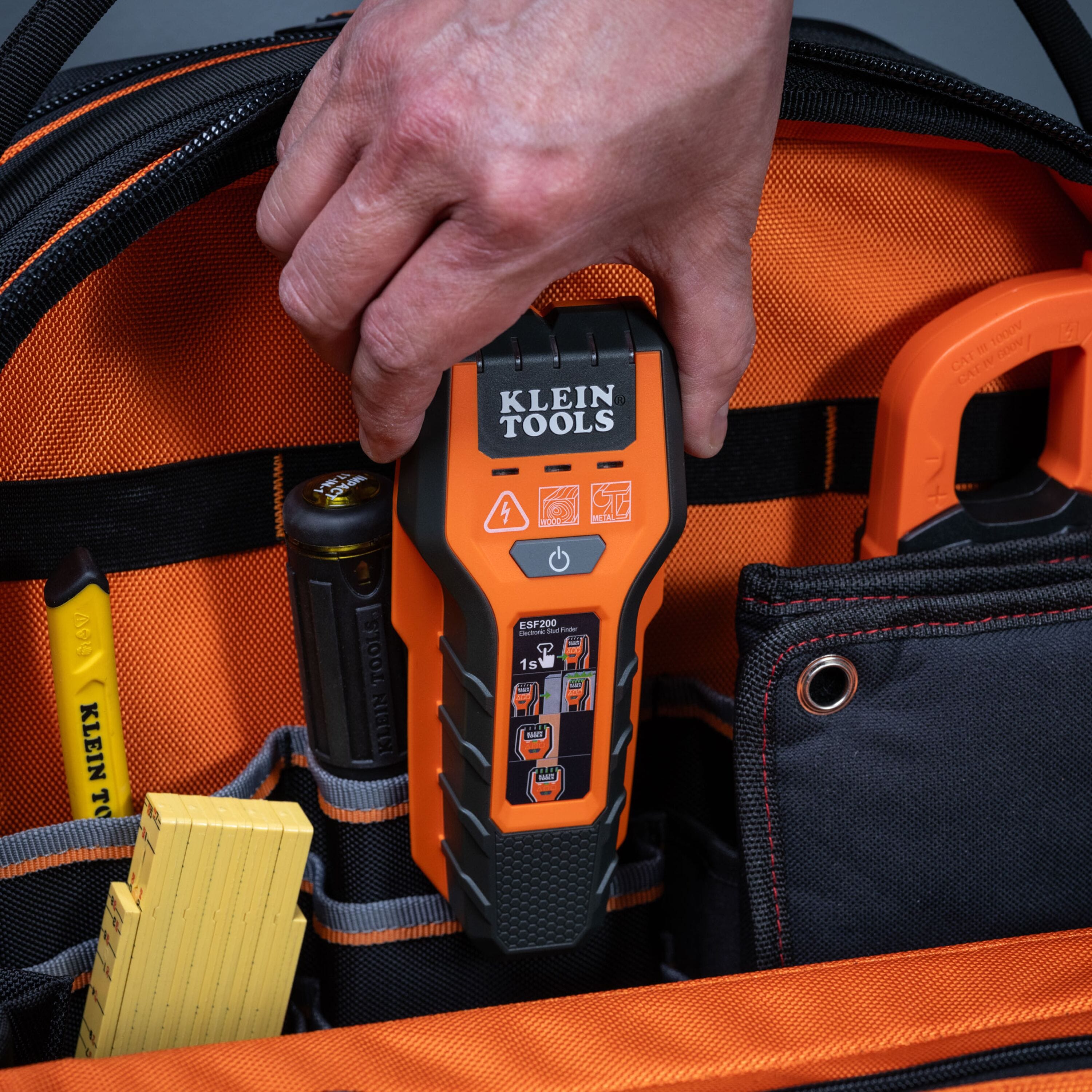 An action shot showing a professional tradesperson’s hand reaching for the compact ESF200 stud finder stored inside a specialized pocket of an orange and black Klein Tools jobsite tool bag.