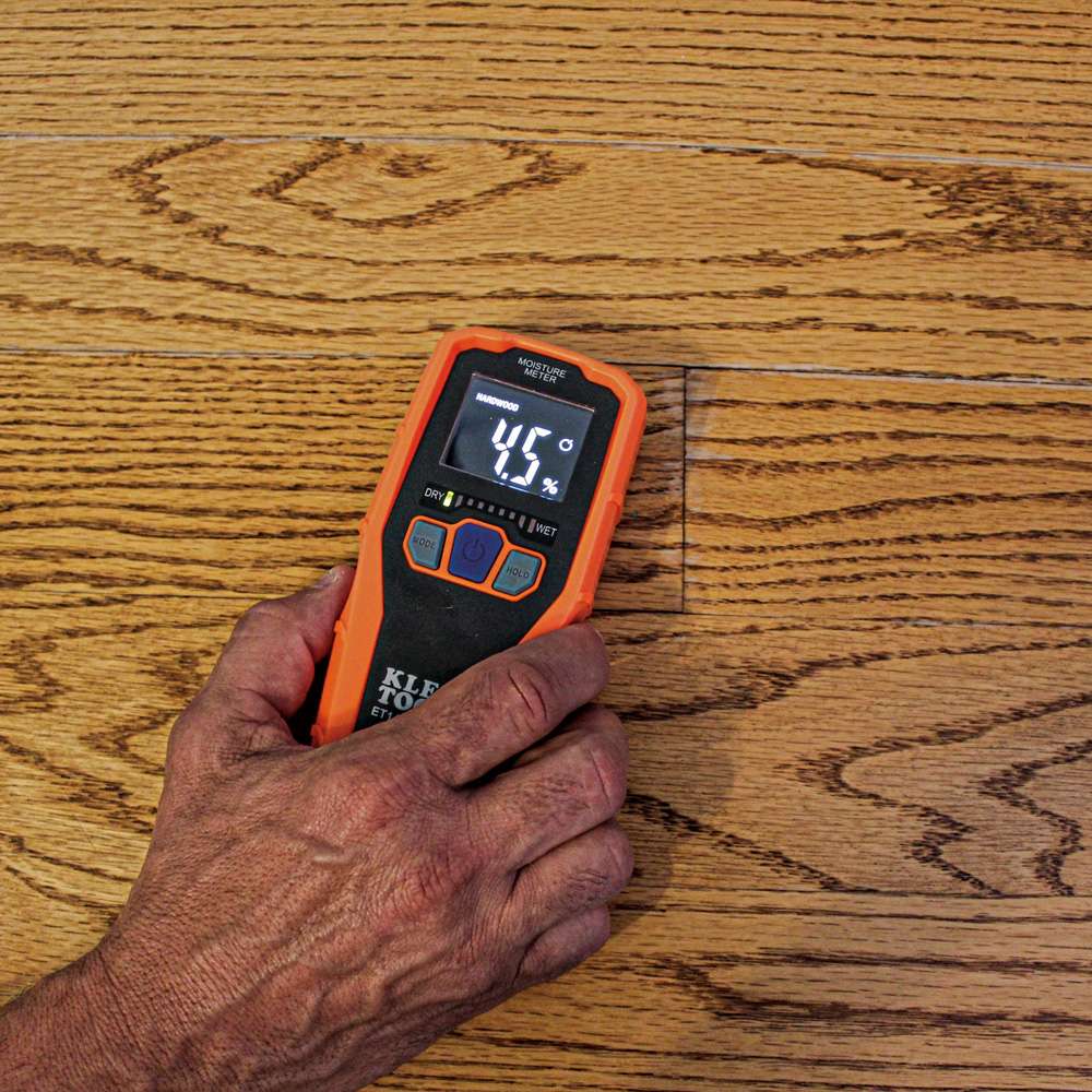 Close-up action shot of a person's hand using the ET140 moisture meter to scan a hardwood floor. The device is set to hardwood mode and displays a relative moisture reading of 4.5 percent with green LED bars on the graph.