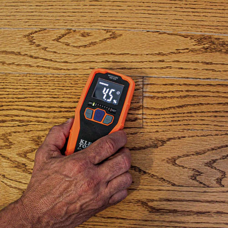 Close-up action shot of a person's hand using the ET140 moisture meter to scan a hardwood floor. The device is set to hardwood mode and displays a relative moisture reading of 4.5 percent with green LED bars on the graph.