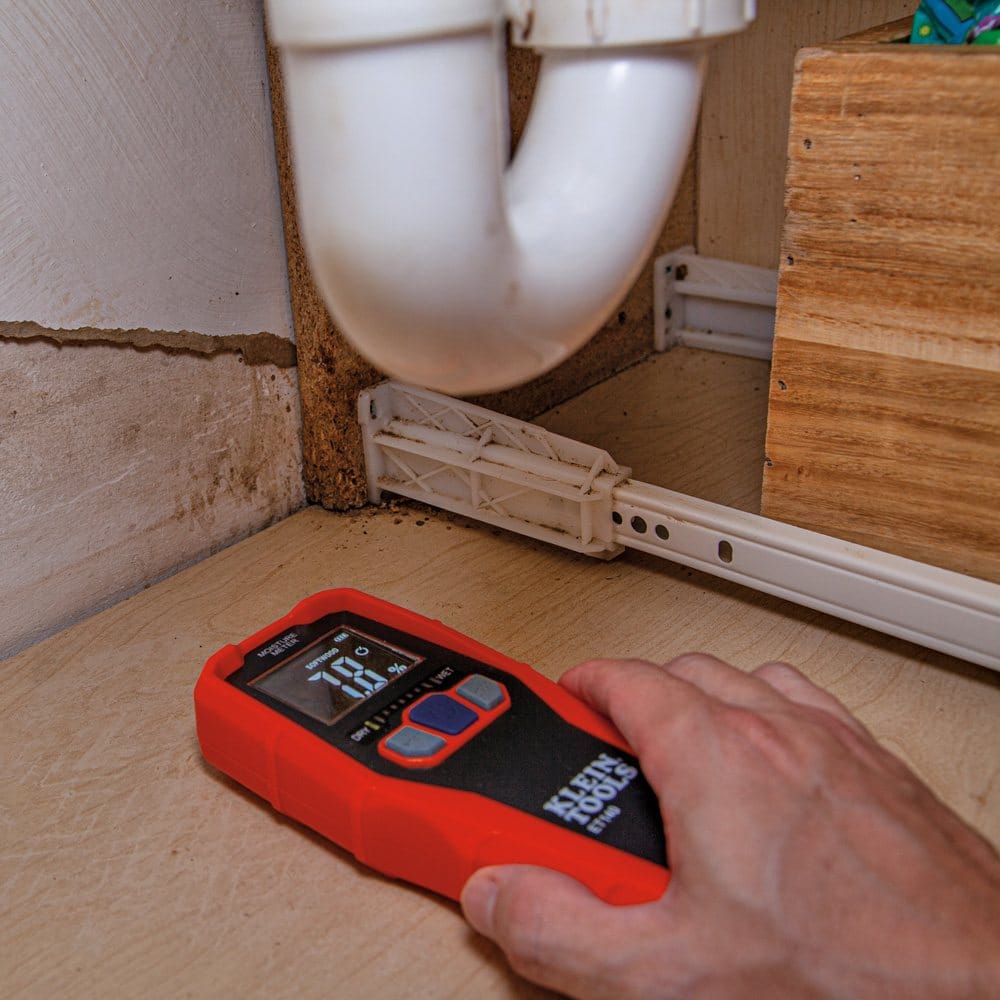 Action shot showing a hand placing the ET140 moisture meter on the base of a wooden cabinet under a sink. The meter displays a relative moisture content of 7.8 percent with green LED bars illuminated on the graph.
